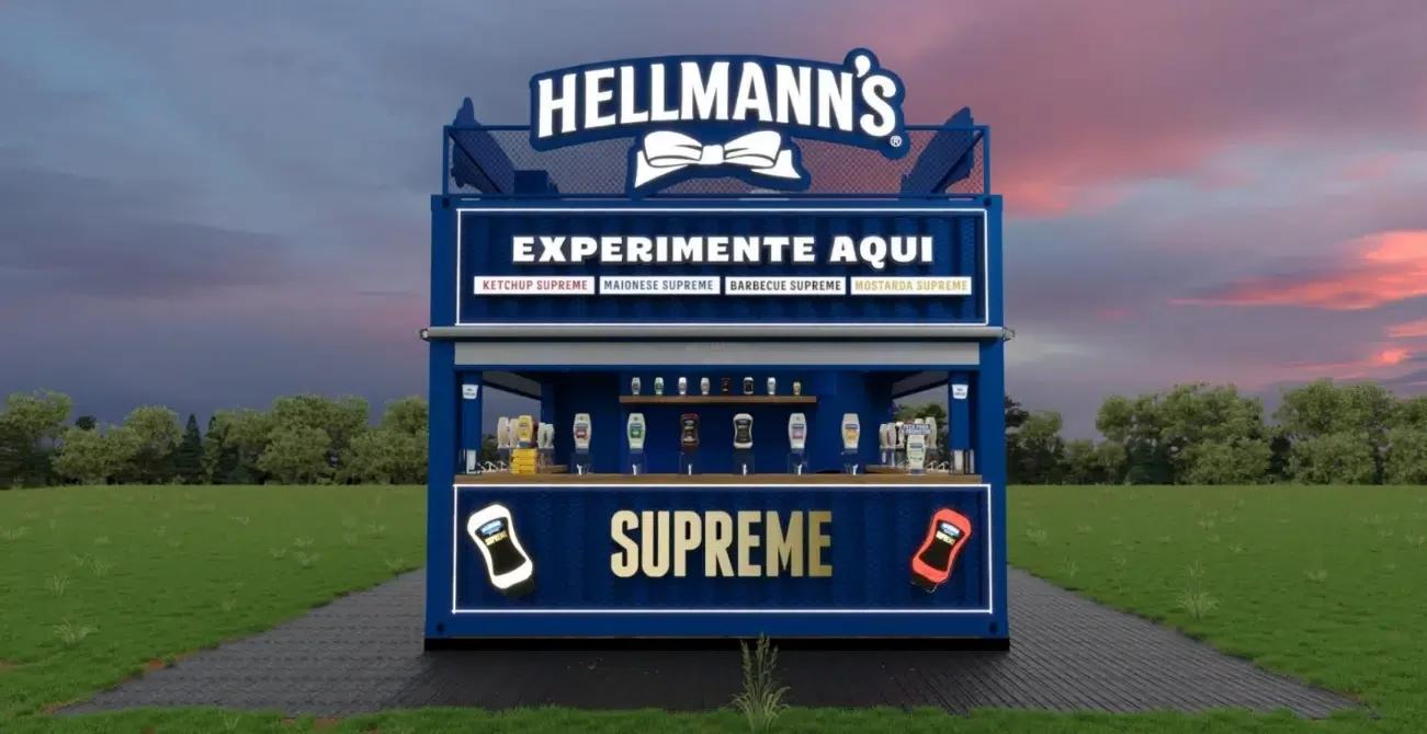 Quiosque azul da Hellmann's feito de contêiner, com letreiro luminoso escrito "Experimente Aqui" e a palavra "Supreme" em destaque na base. O topo exibe o logo da marca sob um céu de entardecer.