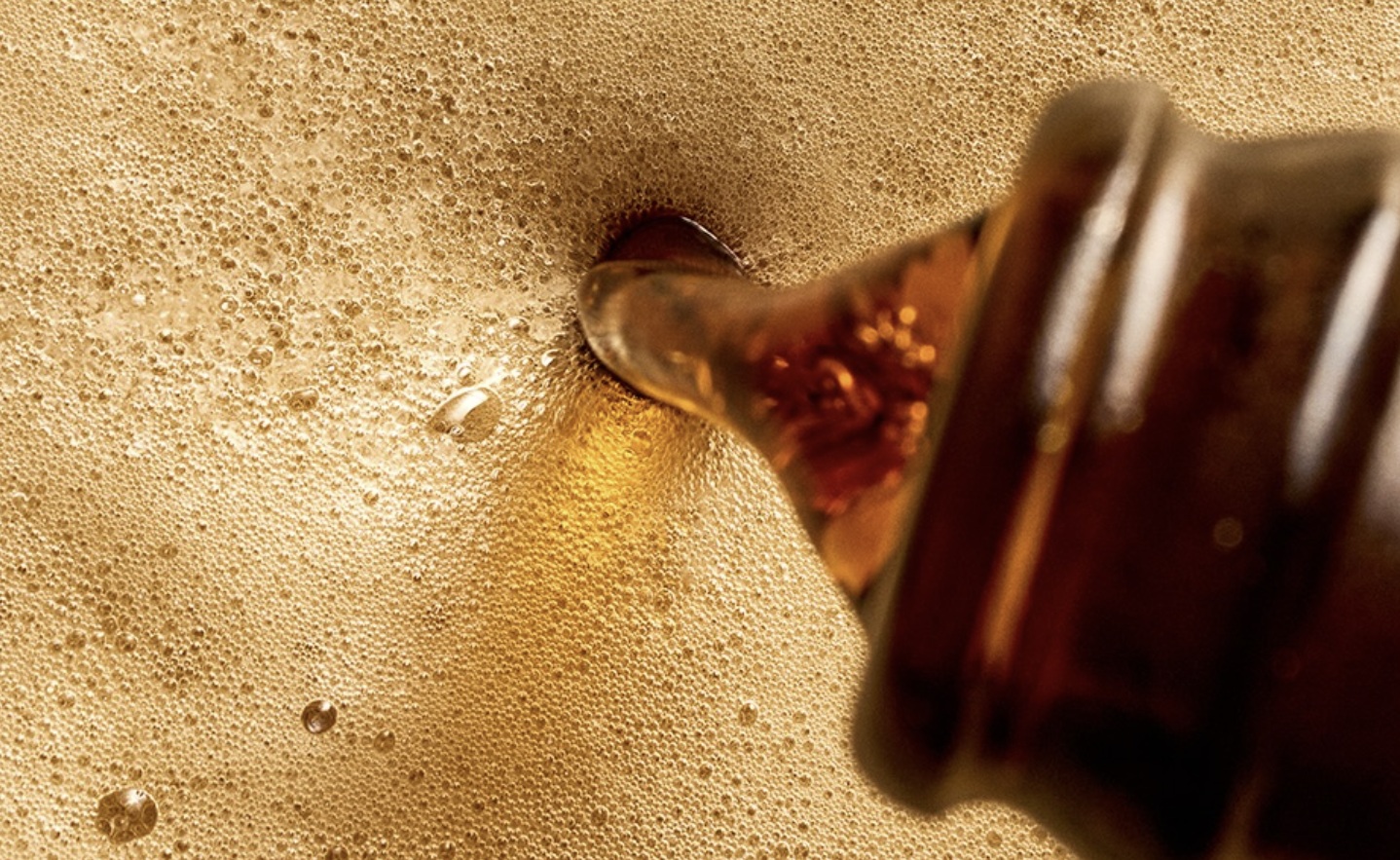 Close macro do bocal de uma garrafa despejando uma cerveja de cor âmbar escura, criando uma camada densa de espuma dourada com muitas bolhas.