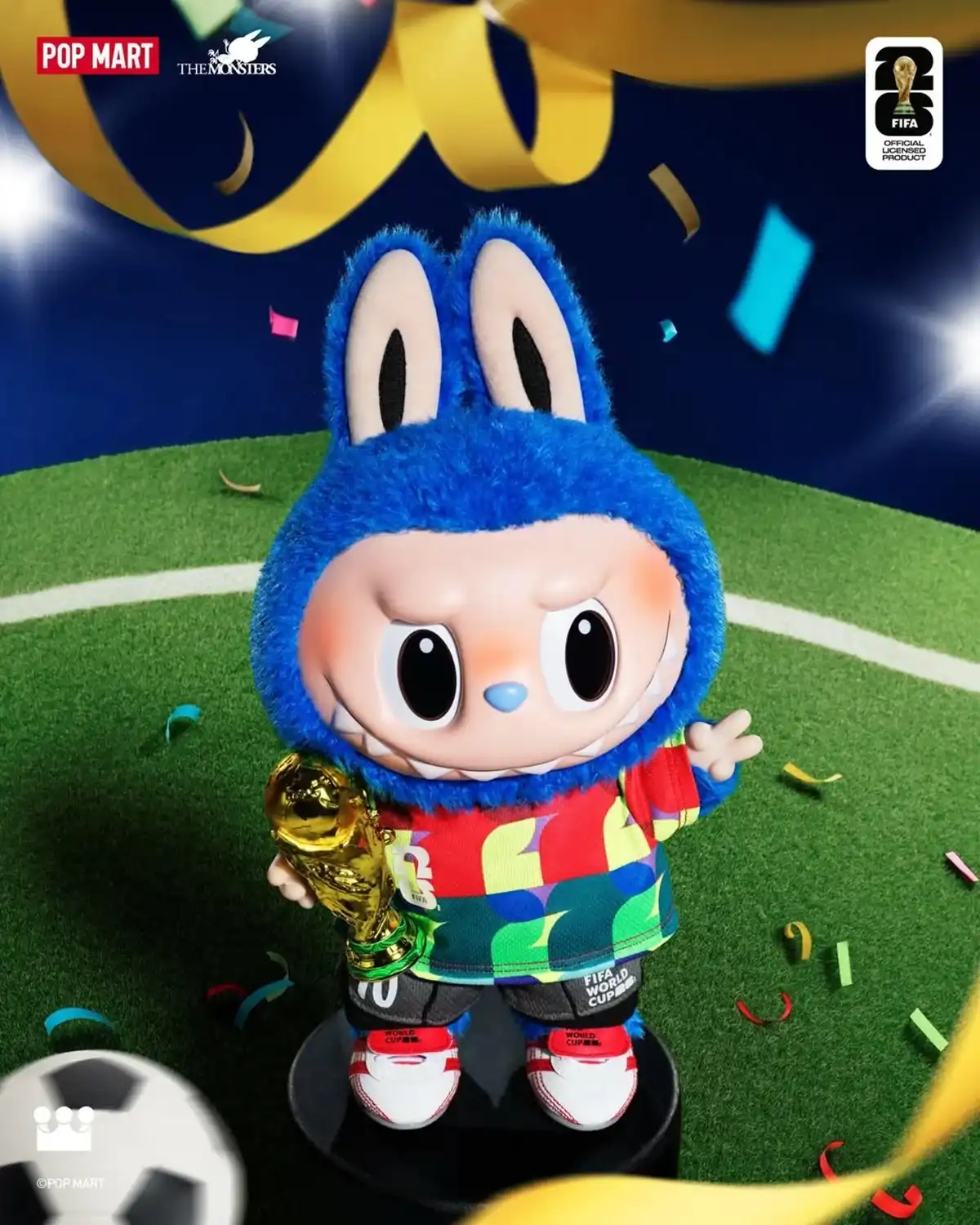 Uma boneca de pelúcia azul e peluda do monstro Labubu, com uma camisa de futebol colorida número 10 e sapatos vermelhos, está em pé em um pequeno suporte preto em um campo de grama verde. Ela está segurando um troféu dourado da Copa do Mundo e acenando com a mão livre. Papel picado colorido cai ao redor dela, e fitas douradas em arco estão no topo da cena em um fundo azul escuro. Os logotipos da marca e da FIFA estão nos cantos superiores.