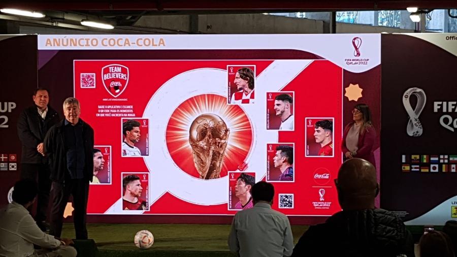 Um grande telão de palco exibe a imagem da página do álbum mostrada na imagem 1, com a taça central e os "Team Believers". Três pessoas estão de pé no palco à esquerda e à direita do telão. Uma plateia está sentada à frente. A borda do telão tem o texto "ANÚNCIO COCA-COLA" e logos da FIFA. Uma bola de futebol está no chão em frente ao palco.