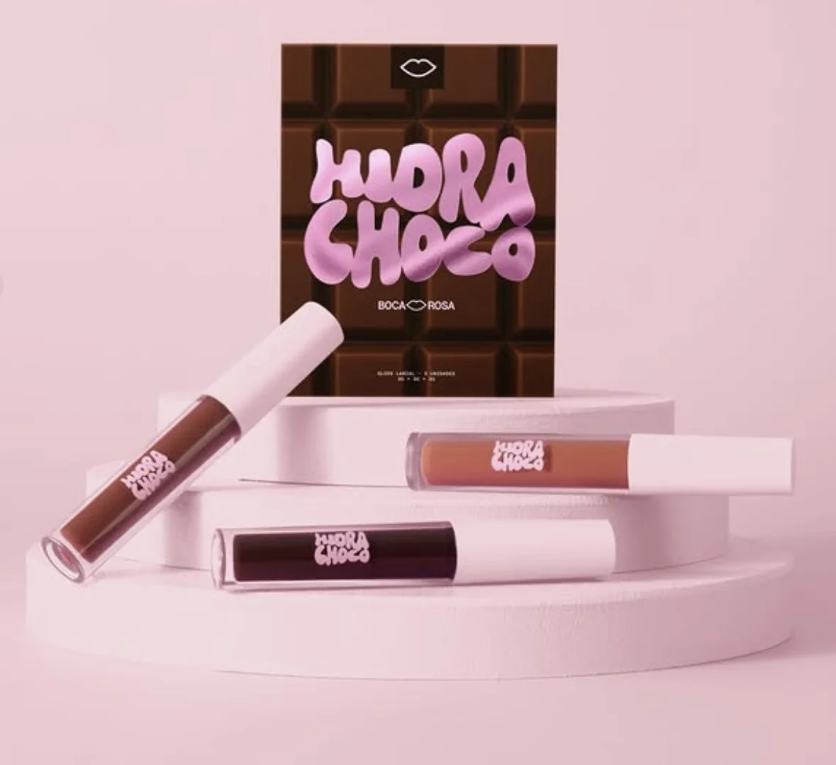 Três embalagens de gloss labial em diferentes tons de marrom dispostas sobre um suporte branco circular. Ao fundo, a caixa do produto imita uma barra de chocolate com a escrita "Hidra Choco".