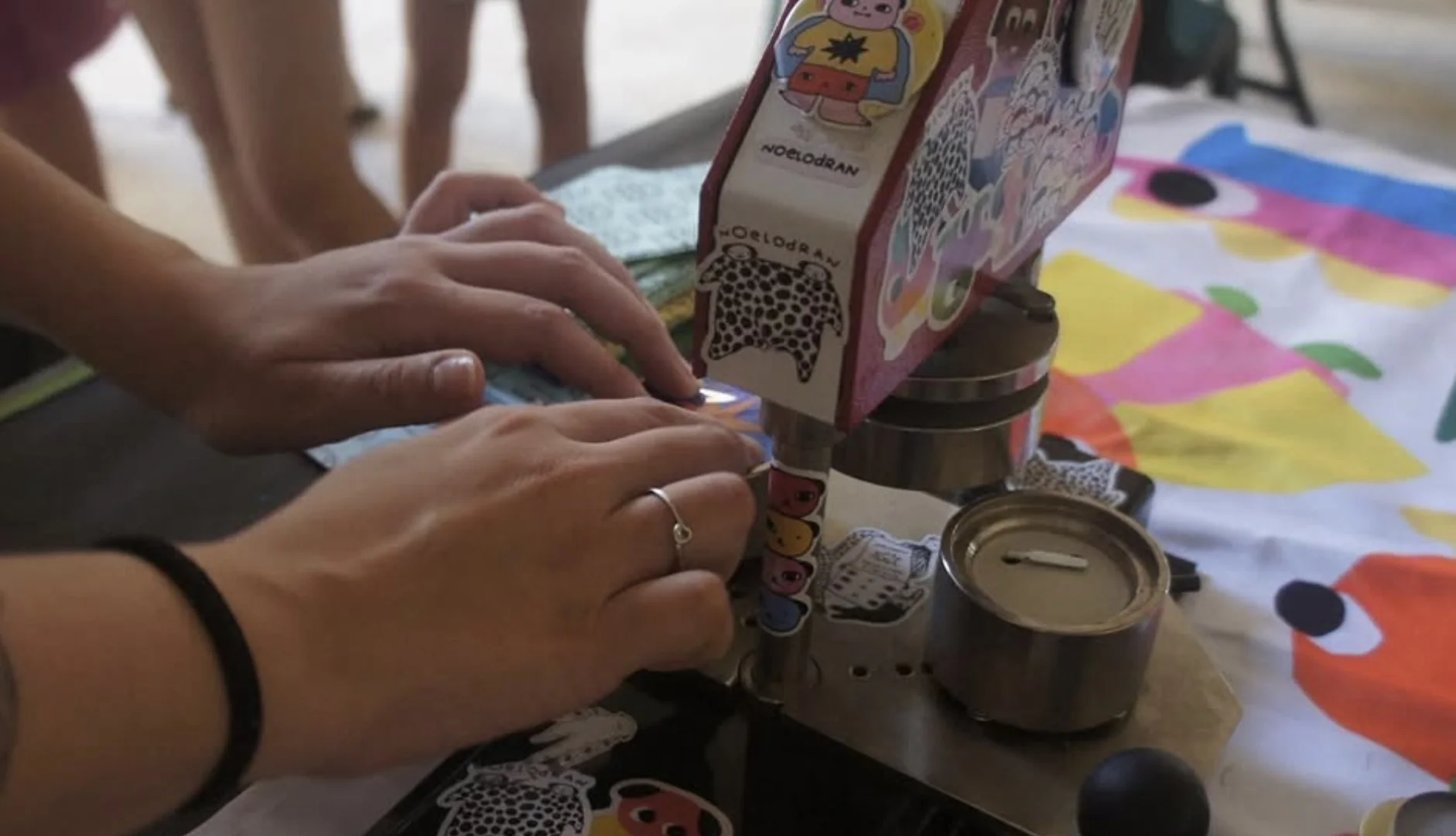 Close-up das mãos de uma pessoa operando uma máquina manual de fazer bottons. A máquina está decorada com diversos adesivos coloridos e ilustrações de personagens.