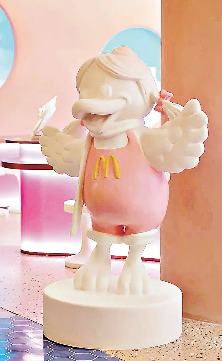 Estátua decorativa da personagem Birdie, do McDonald's, em estilo minimalista branco e rosa, posicionada sobre uma base circular dentro do restaurante temático.