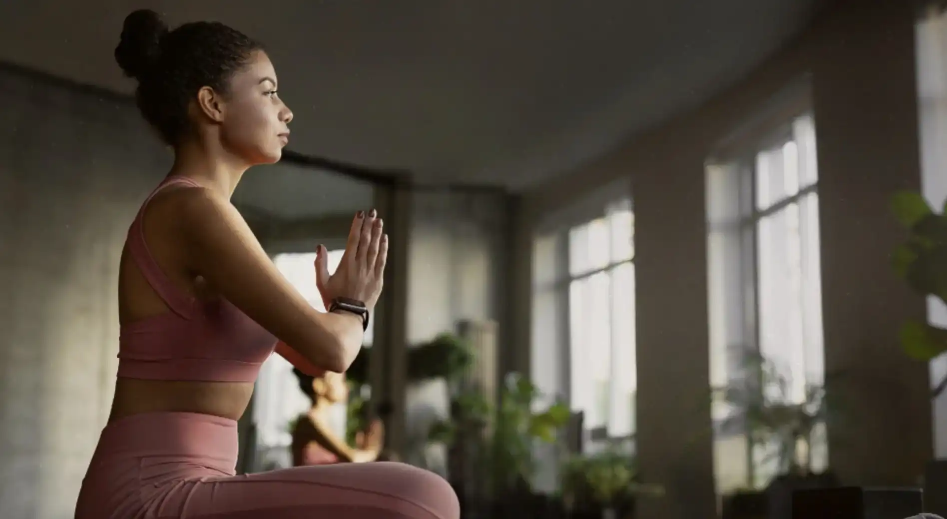 Uma mulher jovem com cabelos presos em um coque, vestindo um conjunto de yoga rosa, sentada em posição de meditação com as mãos unidas em frente ao peito.