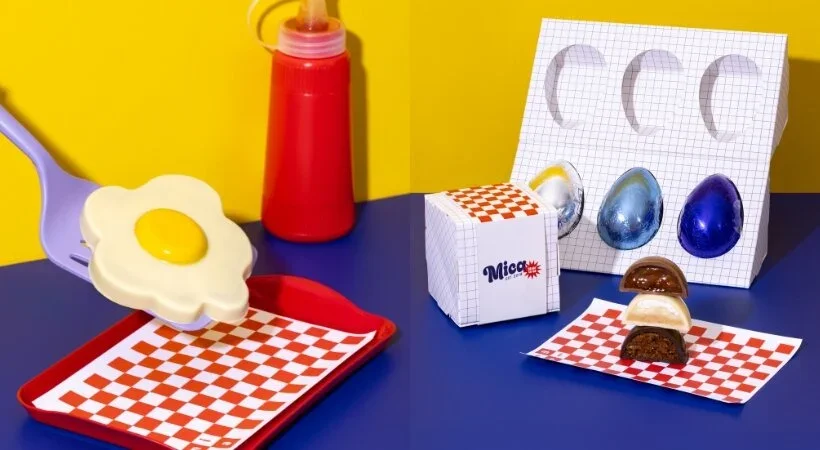 Duas cenas de preparação de chocolate. À esquerda, uma espátula roxa segura um ovo frito feito de chocolate branco e amarelo sobre uma bandeja vermelha com papel xadrez. À direita, um suporte de ovos de papel exibe ovos embrulhados em papel alumínio azul e dourado, ao lado de um mini ovo recheado empilhado. O fundo é uma parede amarela e mesa azul.