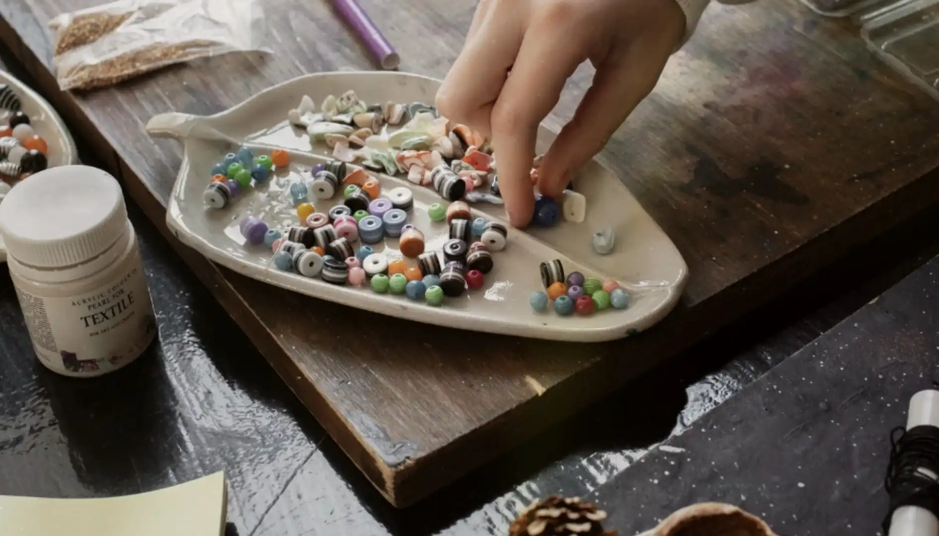 Close das mãos de uma pessoa selecionando miçangas coloridas em um prato de cerâmica branco em formato de folha sobre uma mesa de madeira.
