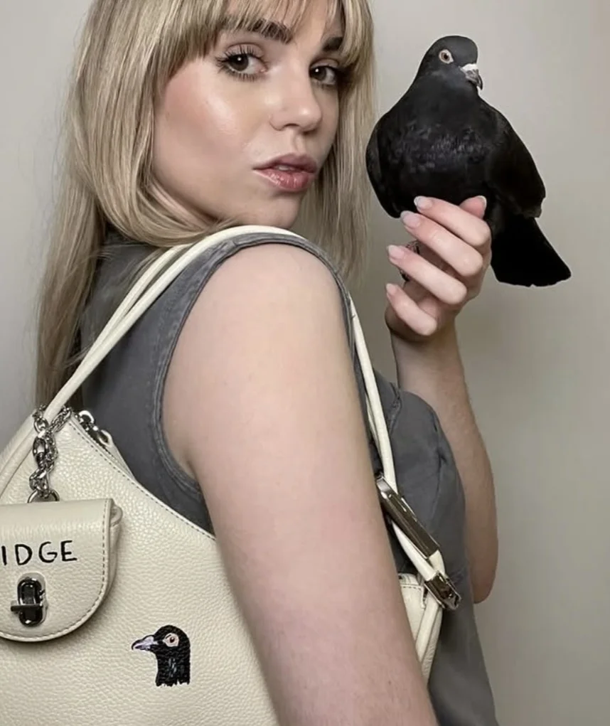 close-up de uma mulher loira com franja olhando para a câmera. Ela segura uma pomba preta na mão e usa uma bolsa de ombro branca com uma ilustração de pomba e o texto "IDGE".