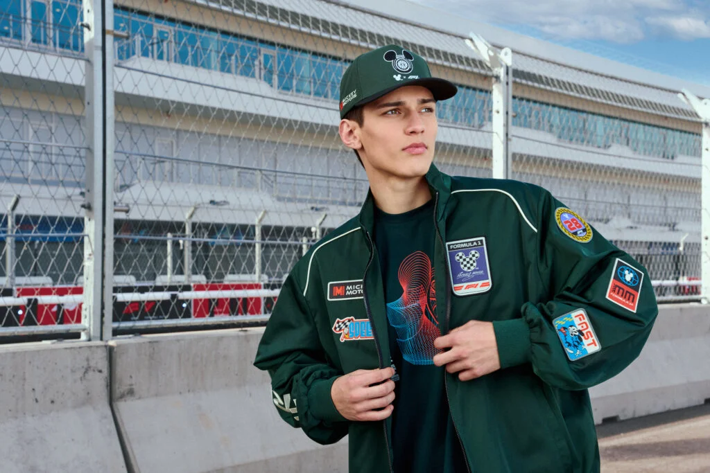 Um jovem modelo posa em um autódromo vestindo uma jaqueta e boné verde-escuro da coleção Disney x Fórmula 1, repletos de patches temáticos do Mickey Mouse e logotipos de corrida.