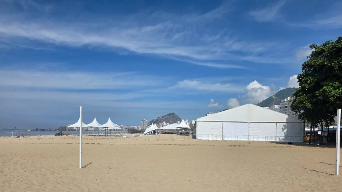 Estruturas de tendas brancas e cercas metálicas sendo montadas na areia da Praia de Copacabana sob um céu azul.