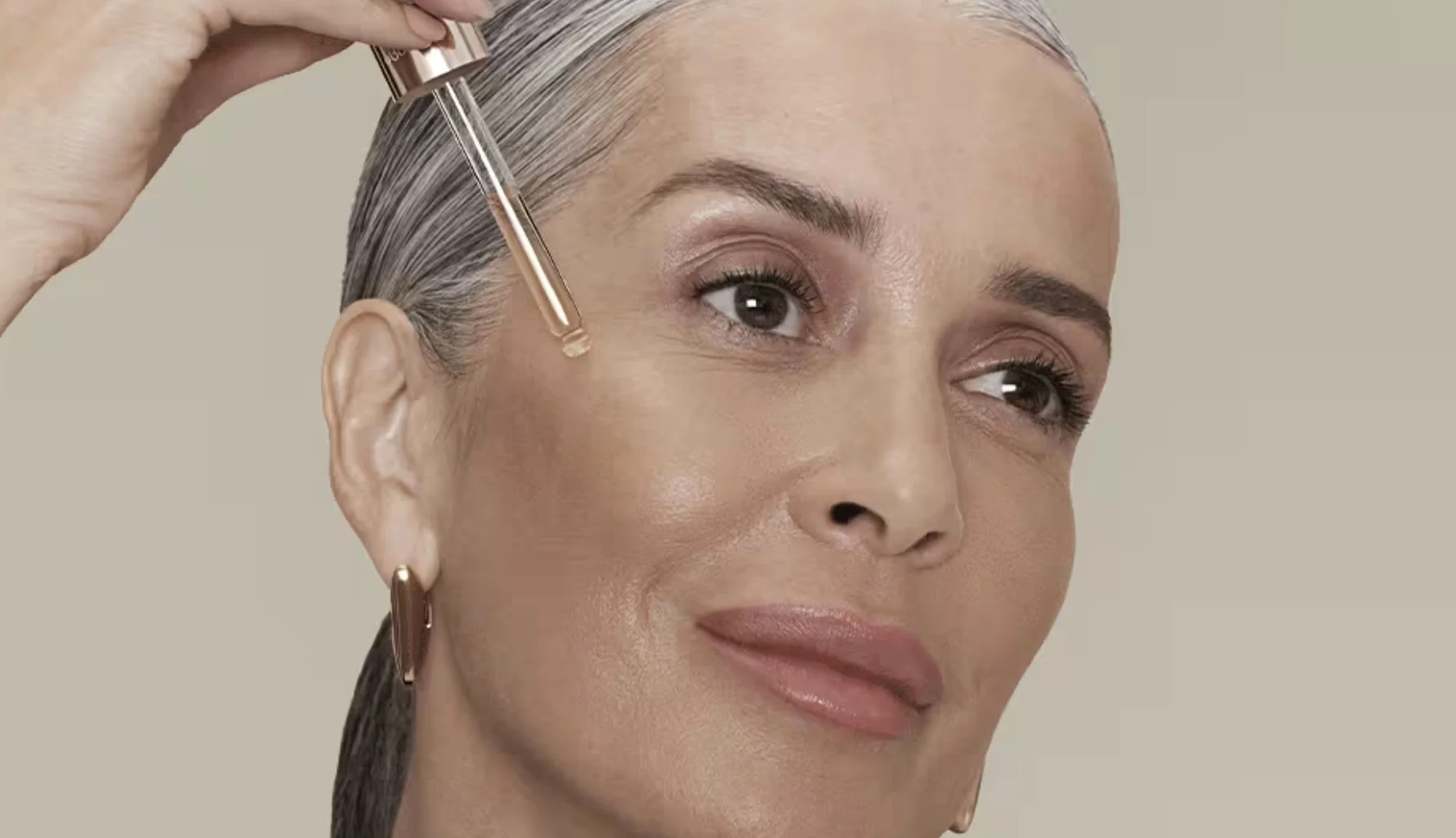 Close-up de uma mulher madura com cabelos grisalhos presos, aplicando uma gota de sérum facial na bochecha com um conta-gotas dourado. Ela sorri levemente e olha para o lado.