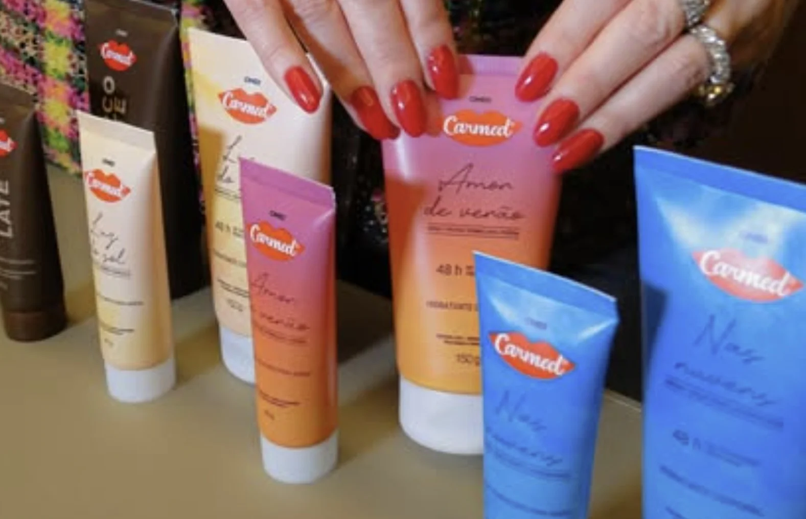 Um close-up focado nas mãos de uma pessoa com unhas vermelhas vibrantes e um anel de diamante em forma de coração, segurando o tubo de 150g do hidratante Carmed Amor de Verão (rosa e laranja) acima de uma mesa com outros tubos desfocados.