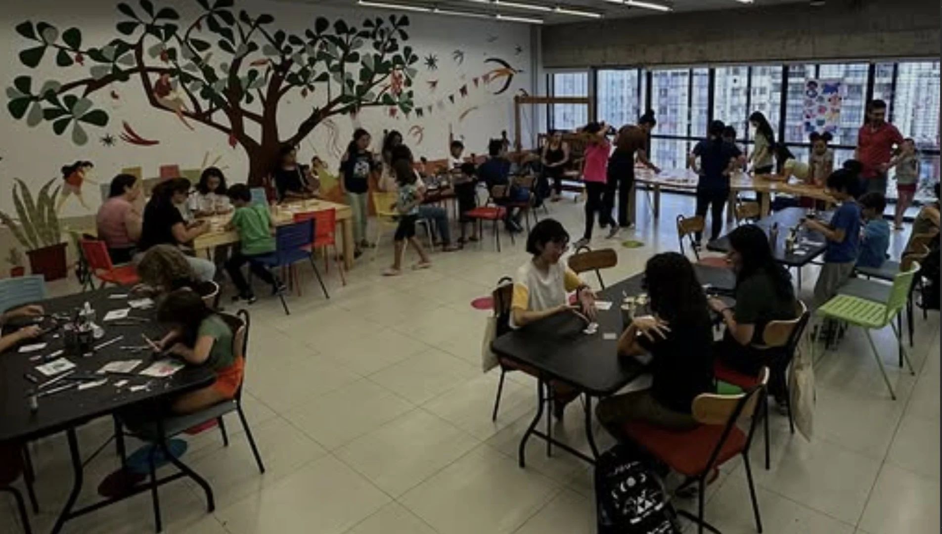 Ampla sala de oficina cultural com crianças e adultos sentados em mesas de trabalho. Ao fundo, uma parede branca exibe um grande mural artístico de uma árvore com folhagens verdes e silhuetas de pessoas.