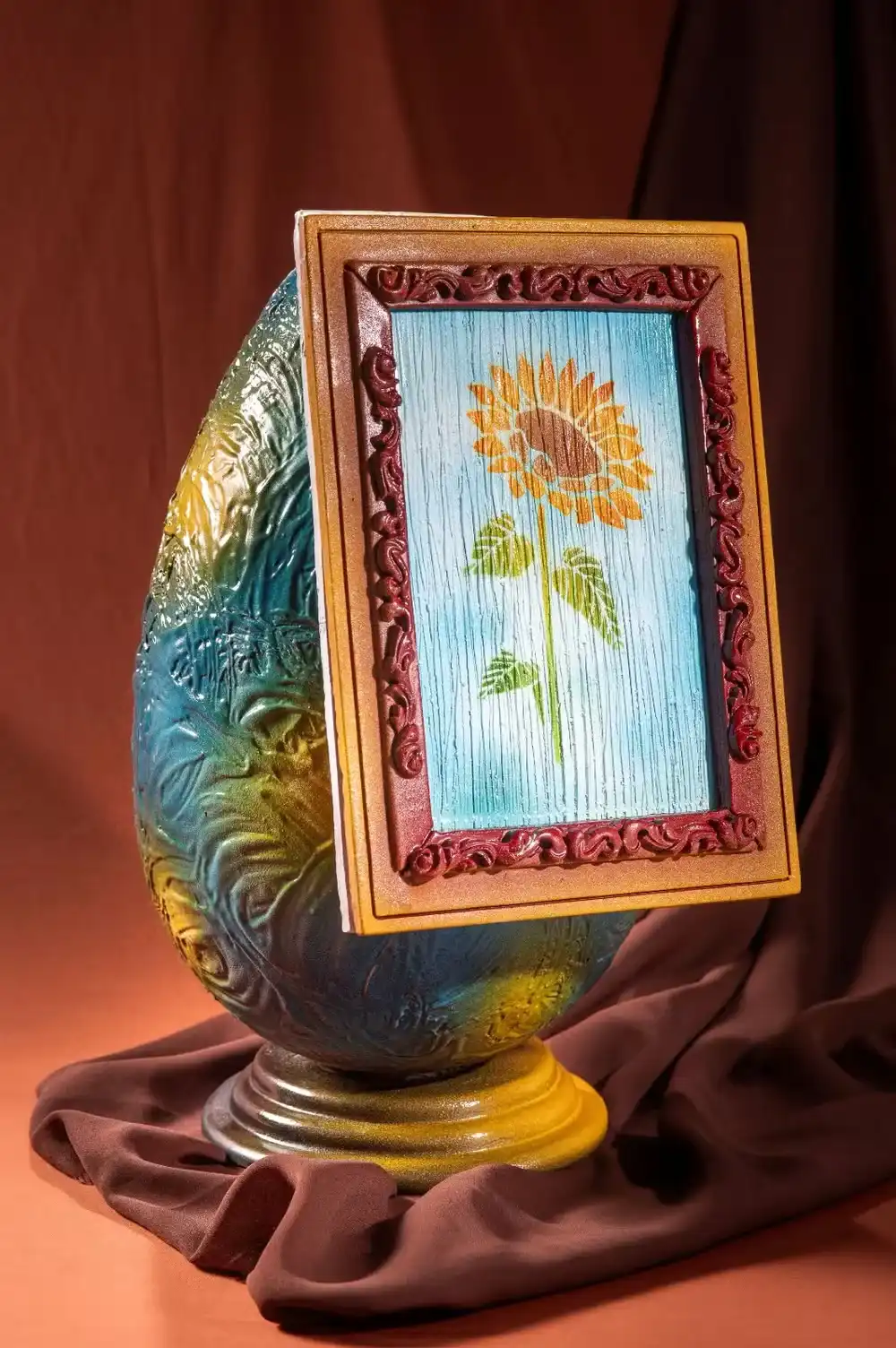 Um ovo decorativo azul e dourado texturizado em um pedestal de latão dourado com um pequeno quadro dourado de um girassol.