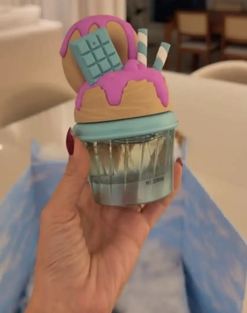 Uma mão segura um frasco de perfume com design criativo que imita um doce ou sobremesa. A base é de vidro transparente facetado, e a tampa grande e detalhada simula uma cobertura de sorvete ou chantilly bege com calda roxa derretida, uma barra de chocolate azul e canudos listrados.