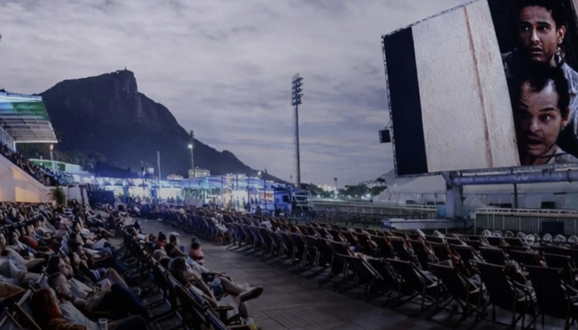 Público sentado em espreguiçadeiras em um cinema ao ar livre à noite, com o Morro do Corcovado ao fundo e o filme "O Auto da Compadecida" na tela.