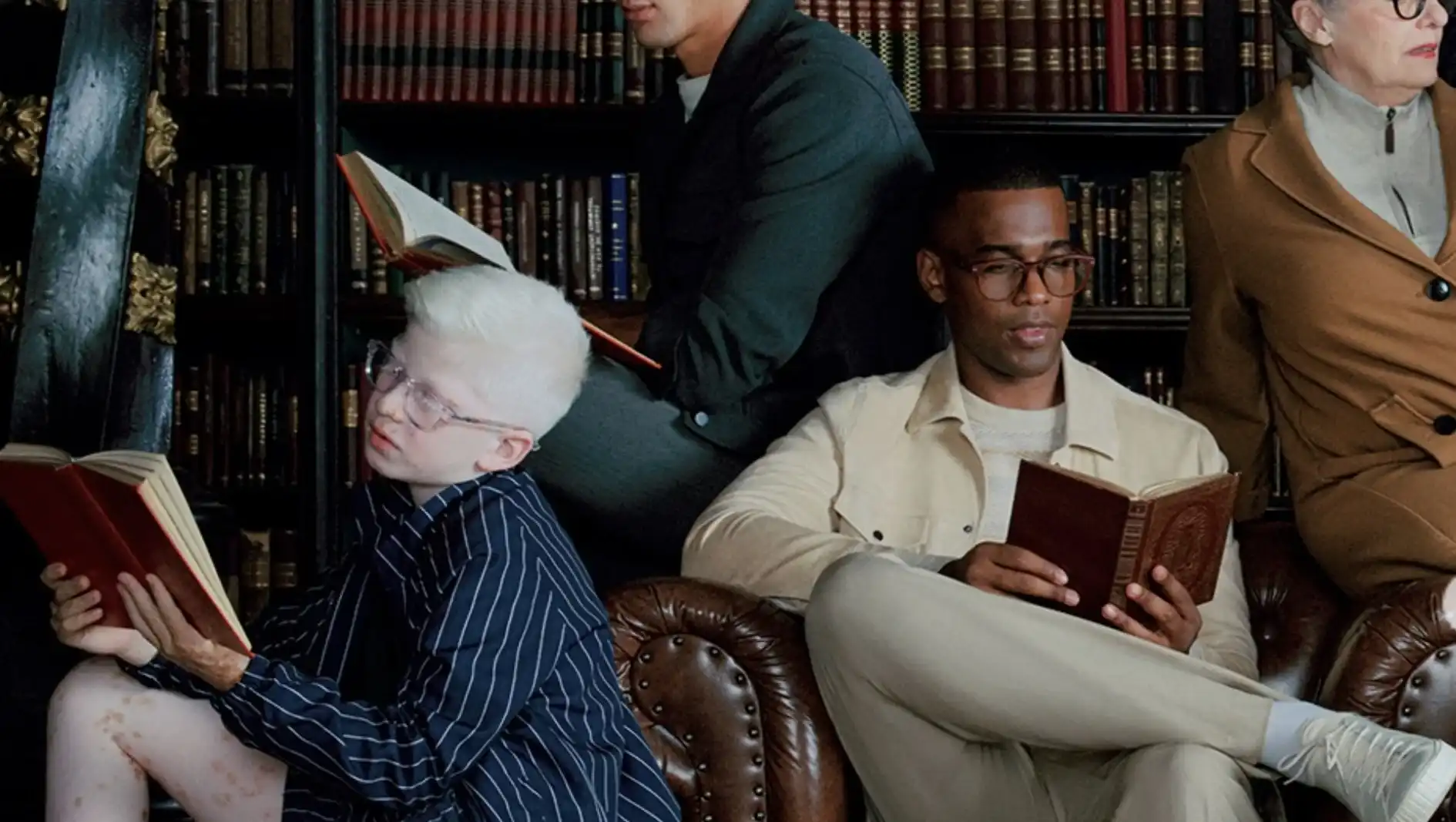 Pessoas de diferentes idades e etnias sentadas em poltronas de couro lendo livros em uma biblioteca clássica com estantes de madeira ao fundo.