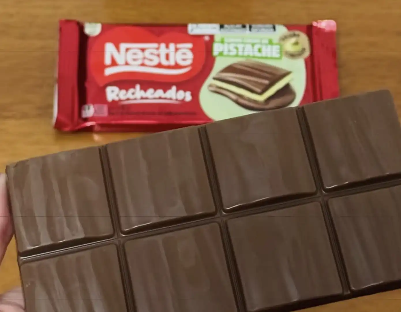 Uma mão segura uma barra de chocolate ao leite aberta em primeiro plano, com a embalagem do Nestlé Recheados sabor Pistache ao fundo sobre uma mesa de madeira.