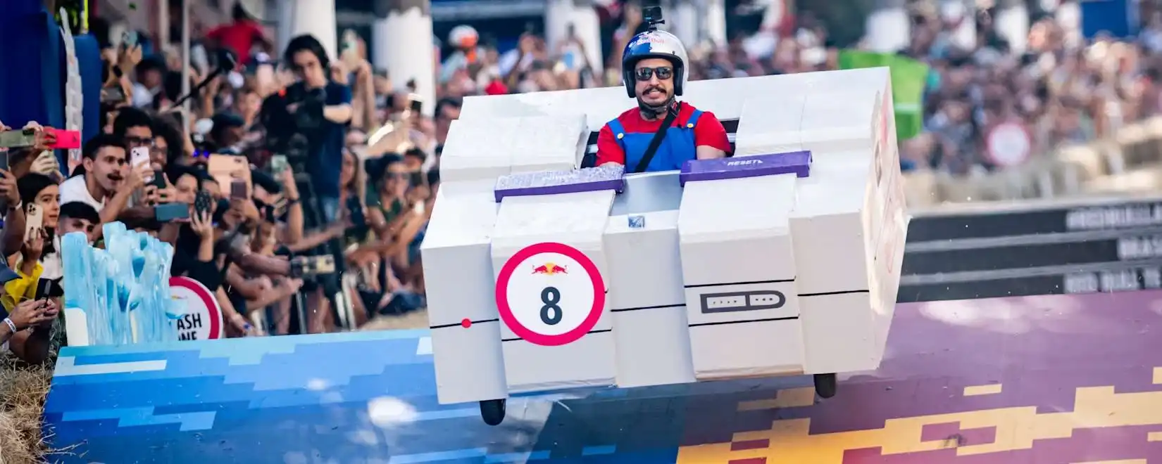 Carrinho de rolimã gigante em formato de console Super Nintendo saltando em uma rampa. O piloto está fantasiado de Mario, usando capacete e óculos escuros, enquanto o público observa ao fundo.