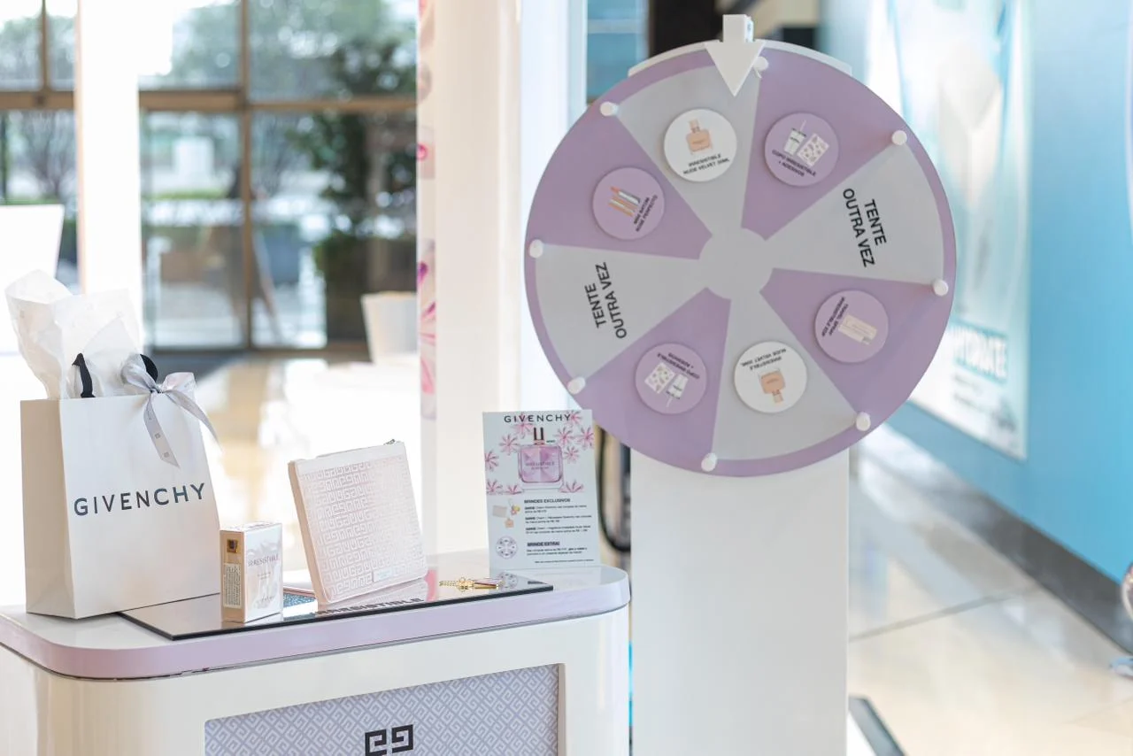 Uma área de brindes e atividades em um evento da Givenchy, com uma roda da fortuna roxa e branca, uma sacola de compras da marca e outros itens promocionais em um balcão.