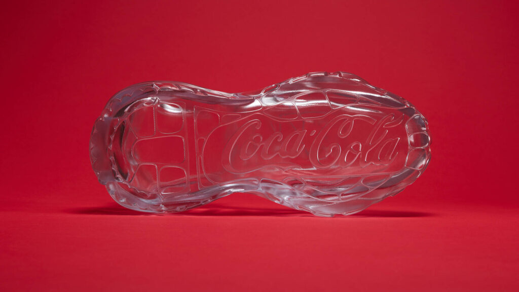 Um close-up em ângulo da sola de vidro de um tênis, mostrando padrões intrincados e o logotipo da Coca-Cola gravado.