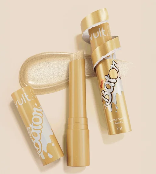 Gloss stick labial da coleção Vult e Baton Garoto na cor pérola/chocolate branco. O produto aparece aberto ao lado de sua embalagem dourada, que simula o papel do chocolate sendo descascado. Ao fundo, uma mancha de textura cremosa com brilho cintilante reforça o acabamento do gloss.