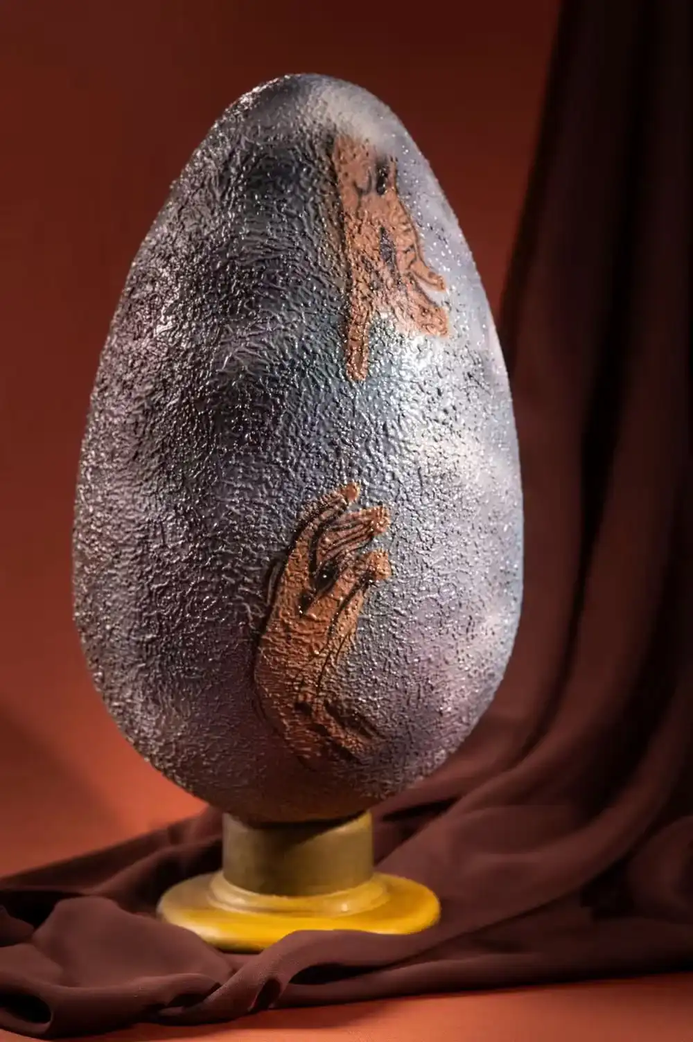 Um ovo decorativo de pedra cinza texturizado em um pedestal de bronze com duas impressões de mãos em cobre.