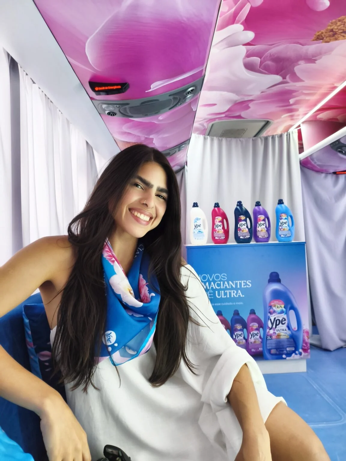 Mulher de cabelos longos e sorriso aberto posa no interior do ônibus da Ypê, usando um lenço azul e branco no pescoço. Ao fundo, um display com cinco frascos de amaciante Ypê Ultra.