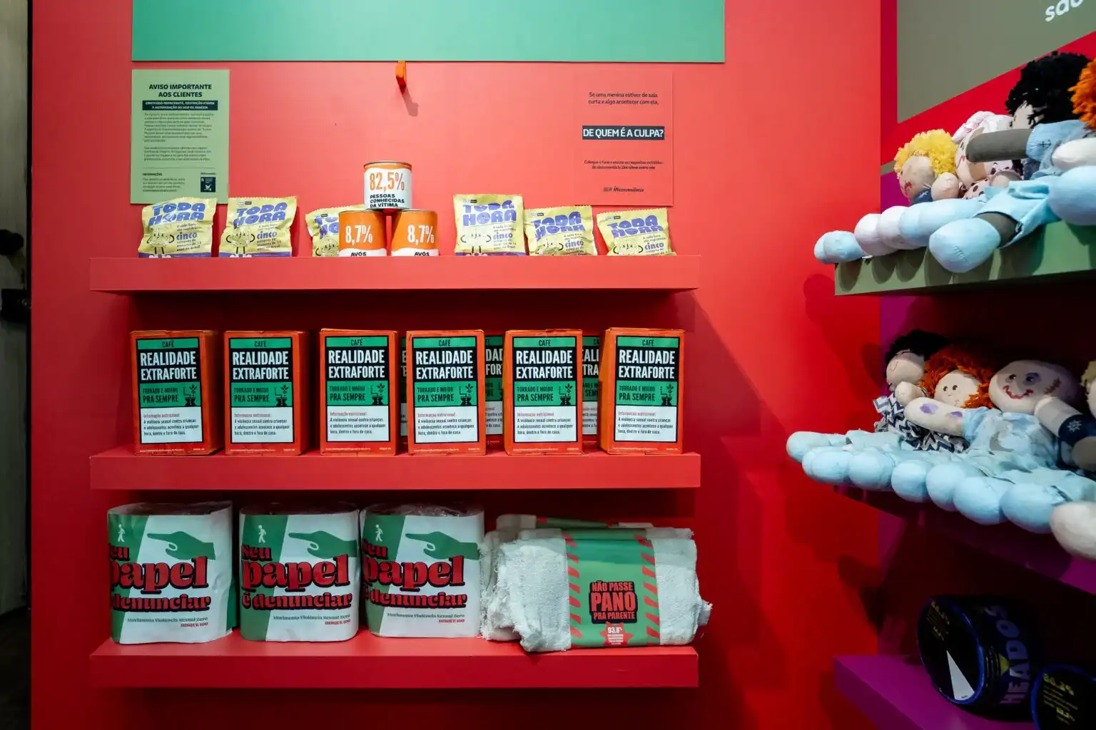 Uma visão mais ampla de uma parede vermelha com três prateleiras flutuantes cheias de produtos de "mercado" com embalagens provocativas. Na prateleira de cima, sacos de "Toda Hora" e latas de "Pessoas conhecidas da vítima" com porcentagens. Na prateleira do meio, várias caixas de café "Realidade Extraforte". Na prateleira de baixo, os rolos de "Seu papel é denunciar" e o pano "Não passe pano". À direita, prateleiras fúcsia contêm bonecas de pano. Placas com texto em português, incluindo "DE QUEM É A CULPA?", estão afixadas na parede vermelha.