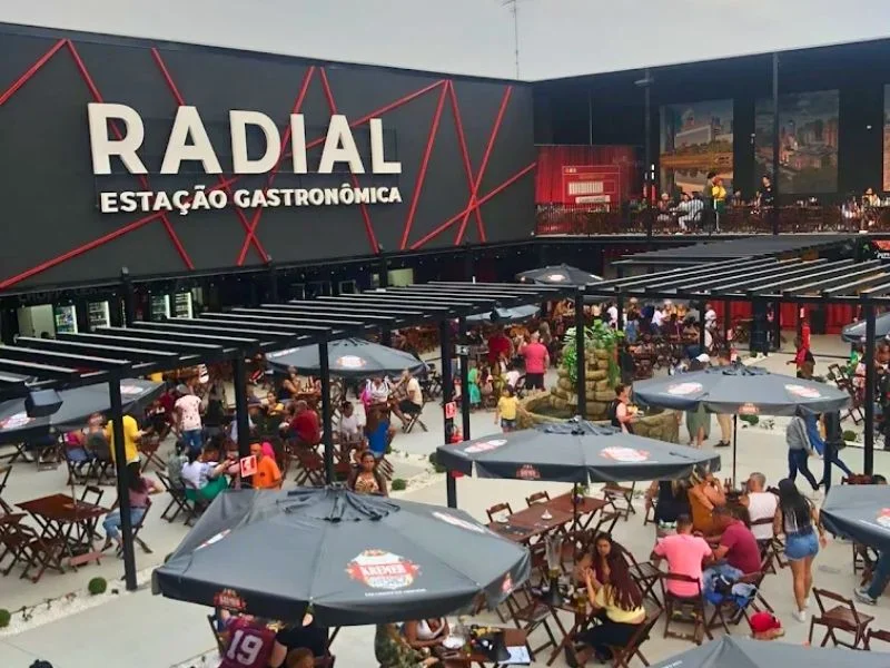 Uma vista aérea de um pátio de praça de alimentação ao ar livre em expansão, com várias mesas, cadeiras e guarda-sóis com marca. A cena está cheia de pessoas jantando e socializando. O edifício principal no fundo tem um letreiro grande e proeminente que diz "RADIAL ESTAÇÃO GASTRONÔMICA".