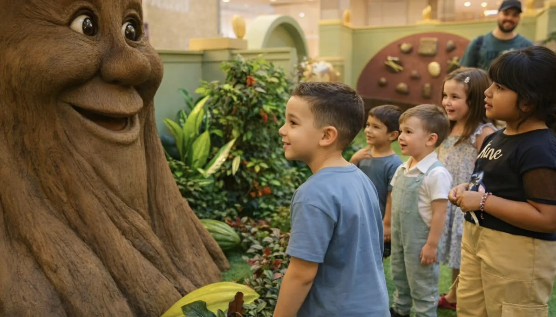 Crianças sorridentes olham para uma árvore falante animatrônica em um cenário de floresta.