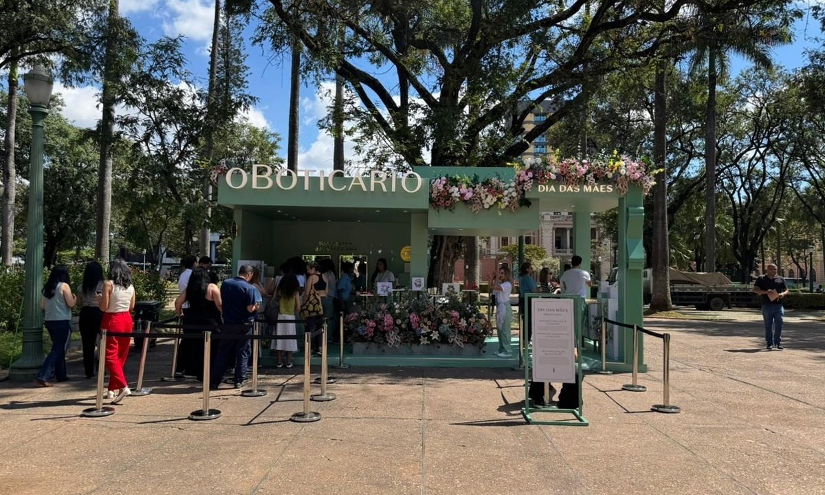 Uma fotografia de uma loja pop-up verde-clara ao ar livre em uma praça com árvores e pessoas na fila. A loja tem a marca "O BOTICÁRIO" em letras brancas na parte superior e um letreiro menor que diz "DIA DAS MÃES". Há arranjos florais exuberantes nas bordas do telhado e perto do chão. Um cordão de segurança organiza uma fila de pessoas à esquerda esperando para entrar.