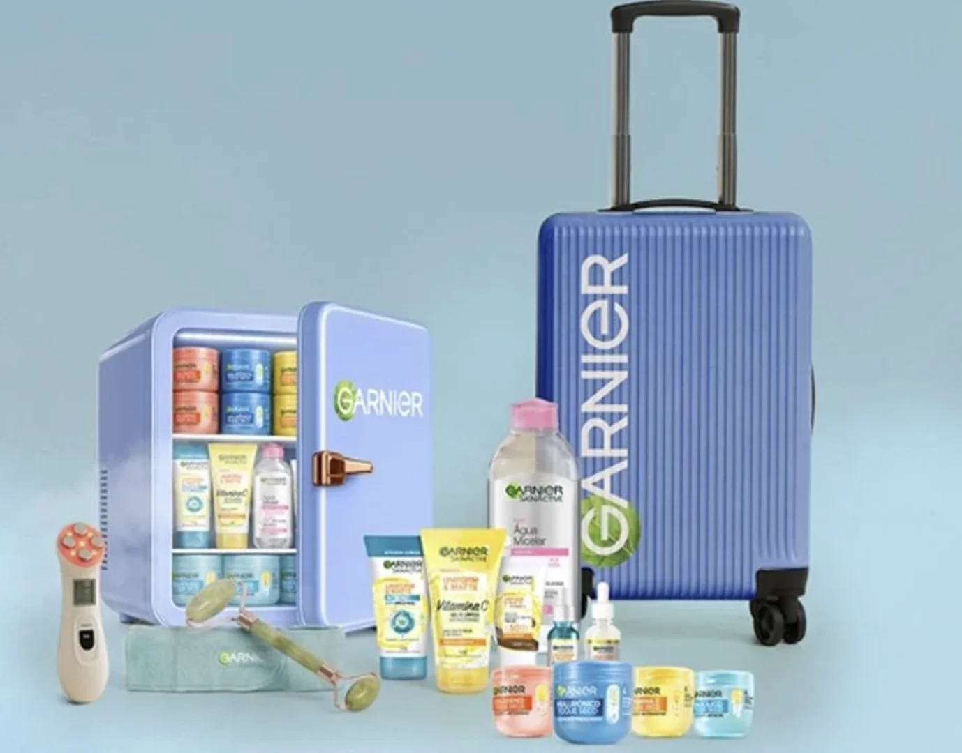 Uma mala de viagem azul e uma mini geladeira lilás com o logotipo da Garnier, cercadas por diversos produtos de cuidados com a pele da marca.