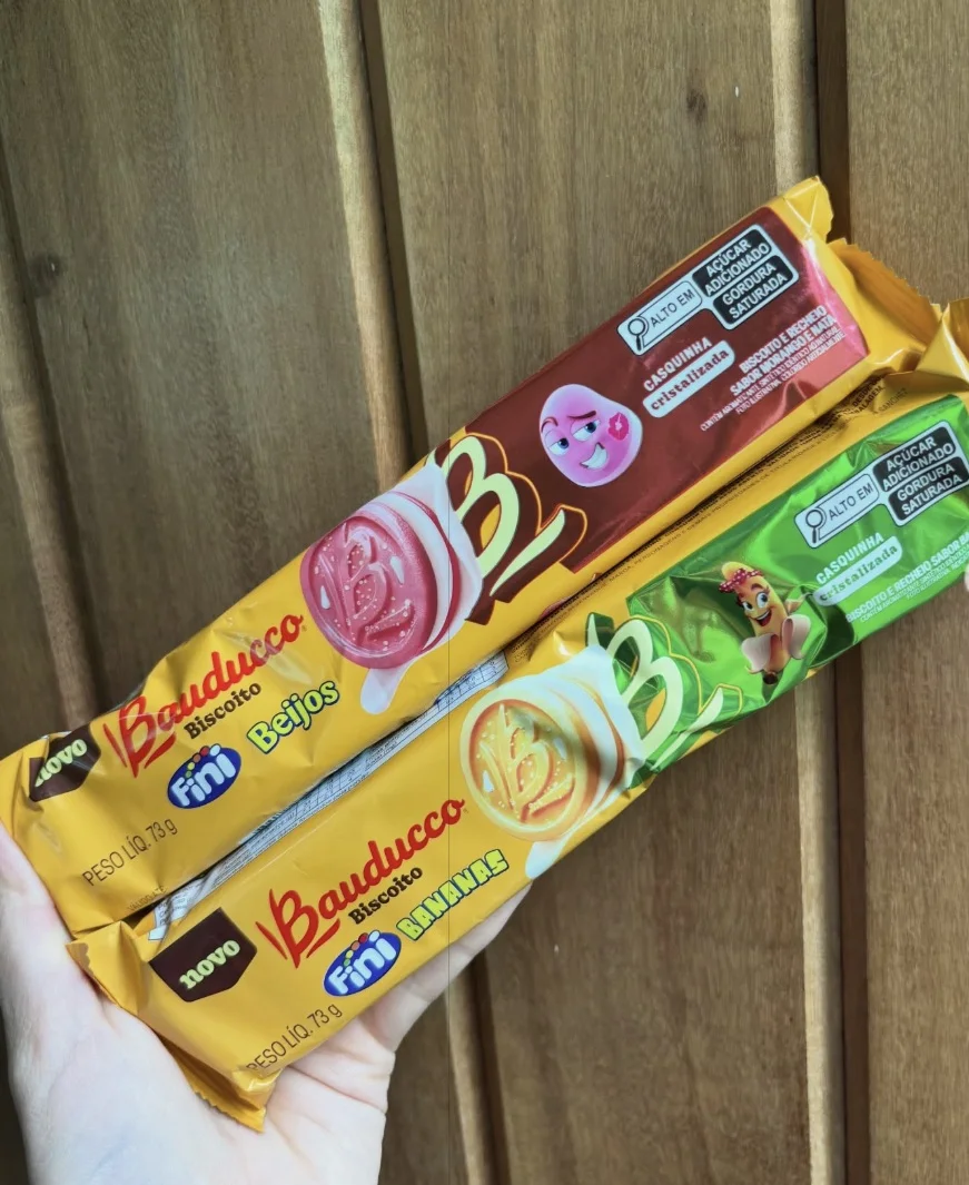 Arte publicitária colorida dos biscoitos Bauducco Fini com os personagens da Fini (Beijo e Banana) e vários biscoitos espalhados sobre um fundo brilhante.