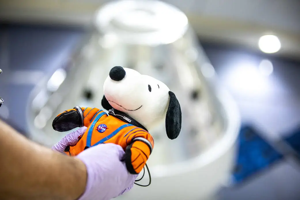 Uma pessoa usando luvas roxas segura um boneco de pelúcia do Snoopy vestido com um traje de astronauta laranja da NASA. Ao fundo, de forma desfocada, aparece uma cápsula espacial metálica.