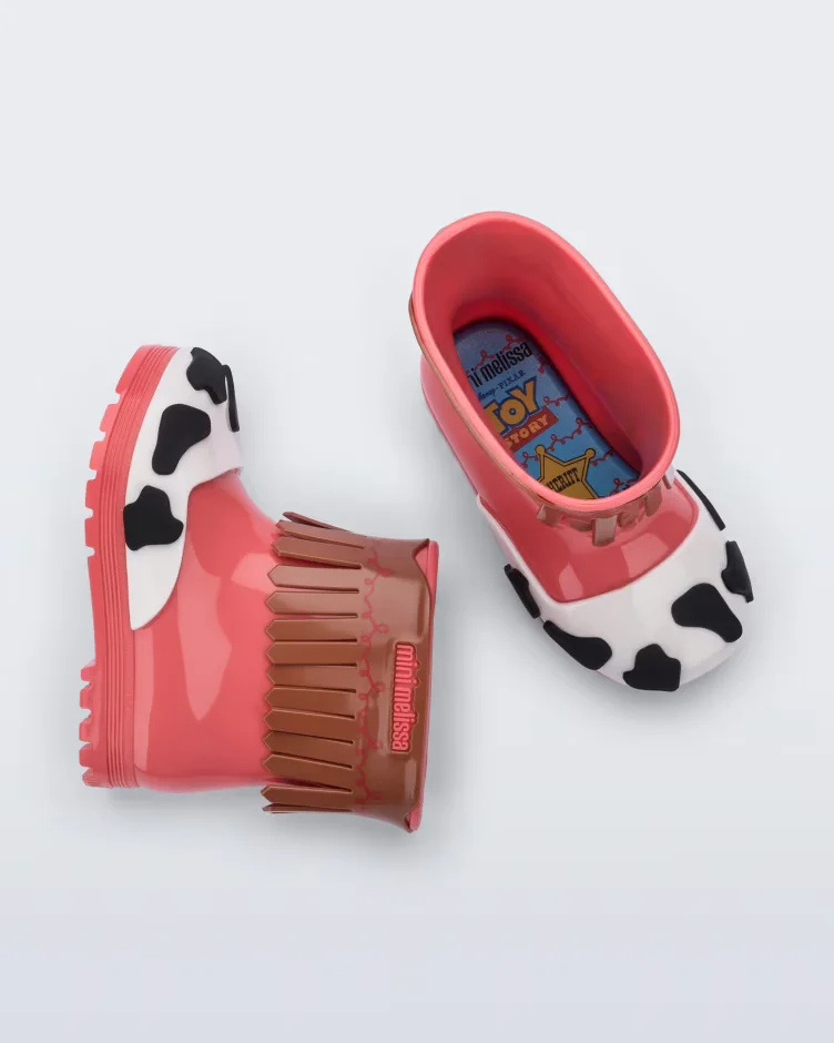 Par de botas infantis de plástico da Mini Melissa, com design inspirado na personagem Jessie de Toy Story, apresentando cores rosa, detalhes de estampa de vaca e franjas marrons.