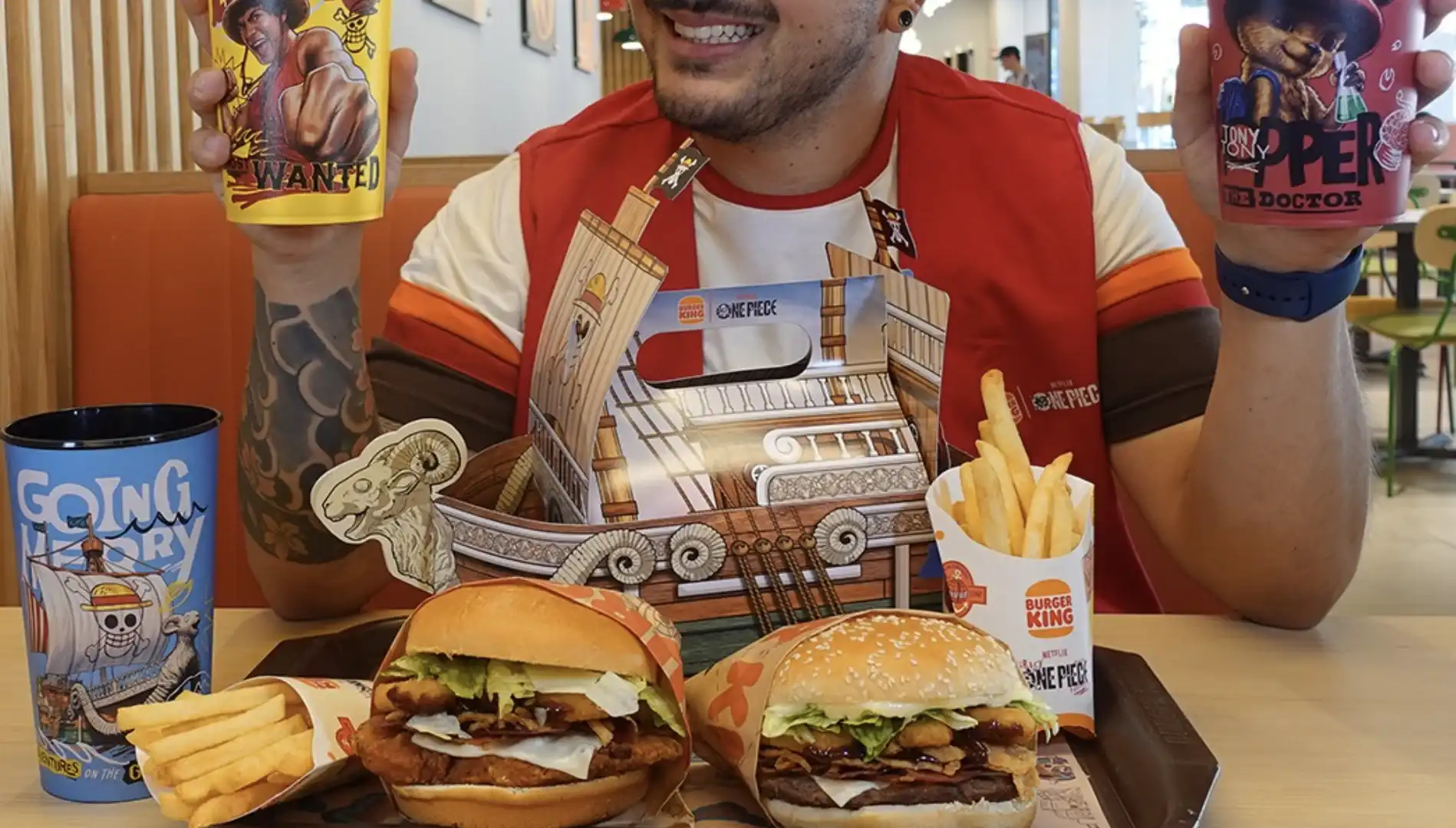 Uma foto de perto, tirada do nível dos olhos, mostra um homem segurando dois copos e um prato com dois hambúrgueres e batatas fritas. O prato também inclui um barco de papelão de "One Piece".