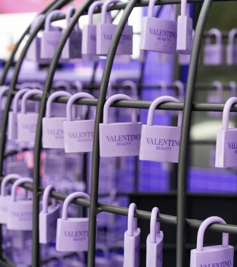 Fileiras de cadeados de latão pintados de lilás fosco pendurados em um rack de metal preto em arco. Cada cadeado tem a inscrição gravada "VALENTINO BEAUTY". O fundo é uma grade de arame azul-púrpura borrada.