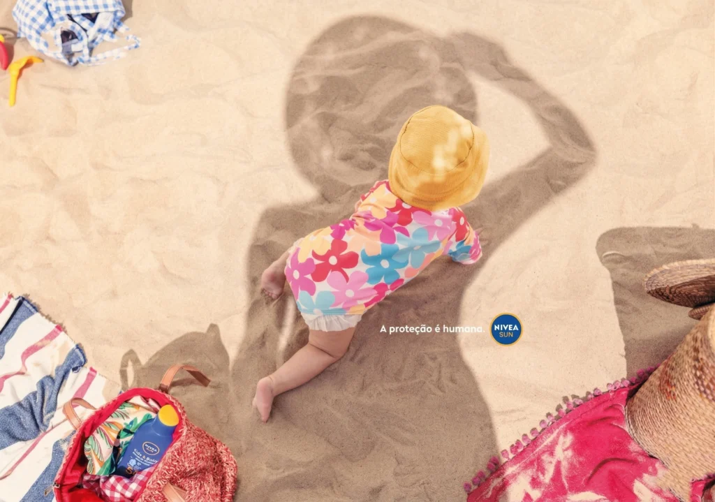 Fotografia em ângulo alto de uma criança engatinhando de costas na areia. Ela veste um maiô floral colorido e um chapéu amarelo. Sua sombra é longa e definida. Há o logotipo da NIVEA SUN e uma bolsa de praia próxima.