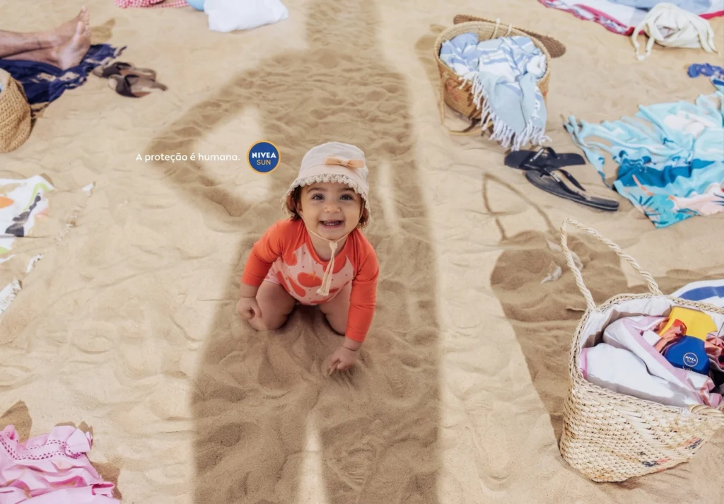 Fotografia vista de cima de uma bebê sorridente na areia da praia. Ela veste um maiô laranja e um chapéu bege de babados. Uma grande sombra protetora a cobre. O logotipo da NIVEA SUN está à direita.
