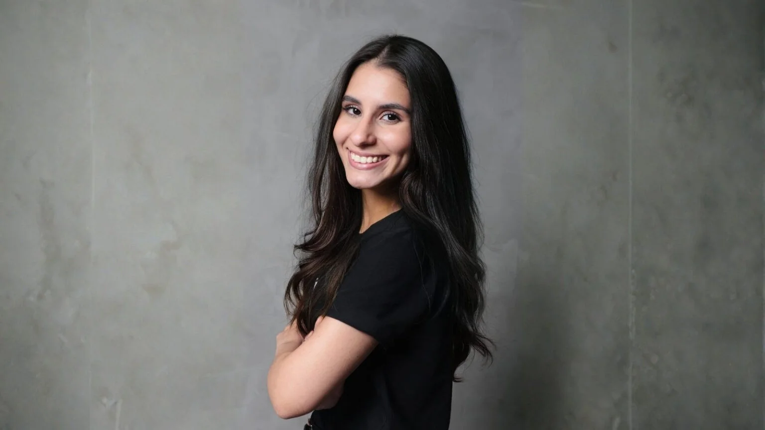 Mulher jovem de cabelos escuros e longos, vestindo camiseta preta, sorrindo de perfil para a câmera contra um fundo cinza neutro.
