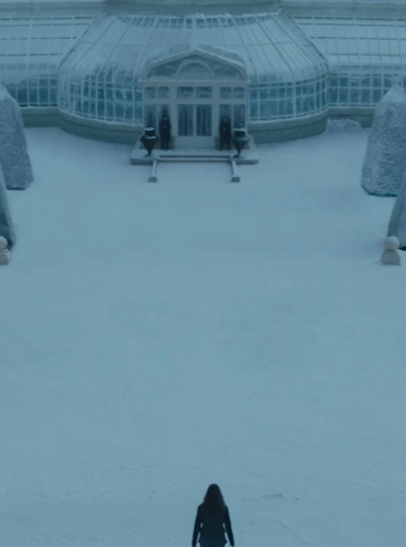 Vista aérea de uma figura solitária de costas, caminhando por um vasto pátio coberto de neve em direção a uma grande estufa de vidro.