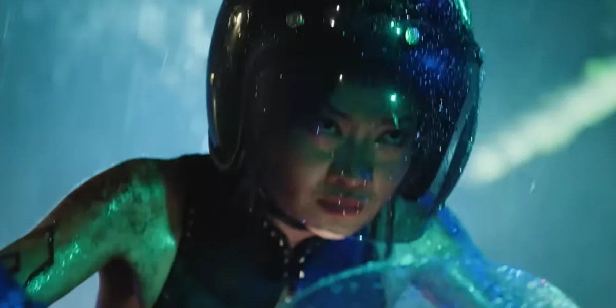 Close-up da personagem Chun-Li usando um capacete de motociclista preto e brilhante sob uma iluminação neon esverdeada e azulada, com gotas de água ou chuva visíveis.