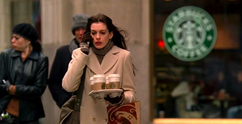 Anne Hathaway como Andy Sachs em "O Diabo Veste Prada", caminhando apressada por uma rua de Nova York enquanto equilibra uma bandeja com quatro copos de café do Starbucks e fala ao celular.