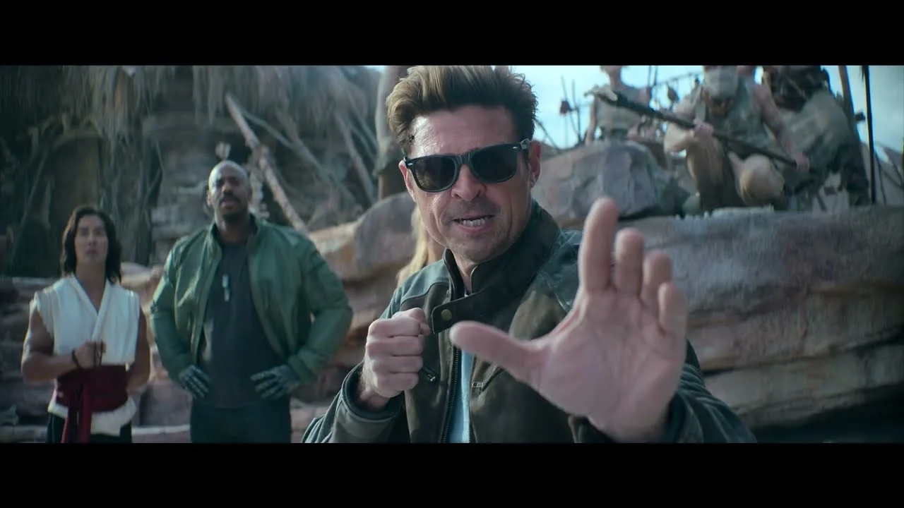 Johnny Cage, interpretado por Karl Urban, em posição de combate usando óculos escuros. Ao fundo, Jax e Liu Kang observam em um cenário rochoso.
