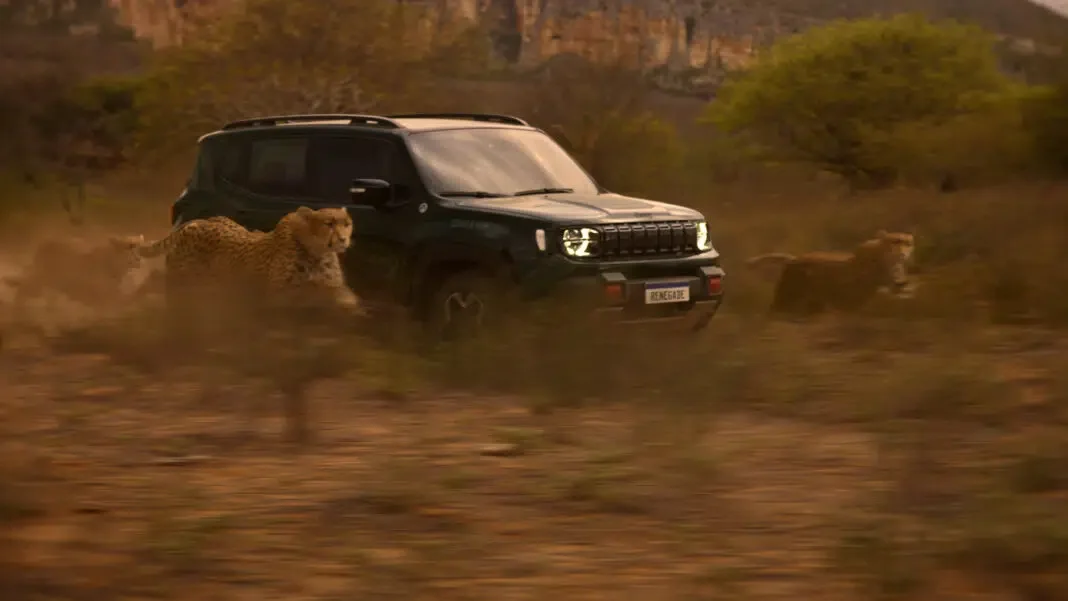 Um SUV Jeep Renegade escuro e robusto percorrendo em alta velocidade uma savana empoeirada e árida, flanqueado e em concorrência direta com dois guepardos em plena corrida. O veículo está ligeiramente à frente de um guepardo à direita e atrás de outro à esquerda, com poeira no ar. A placa frontal diz "RENEGADE". Ao fundo, formações rochosas e vegetação esparsa sob uma luz dourada de pôr do sol.