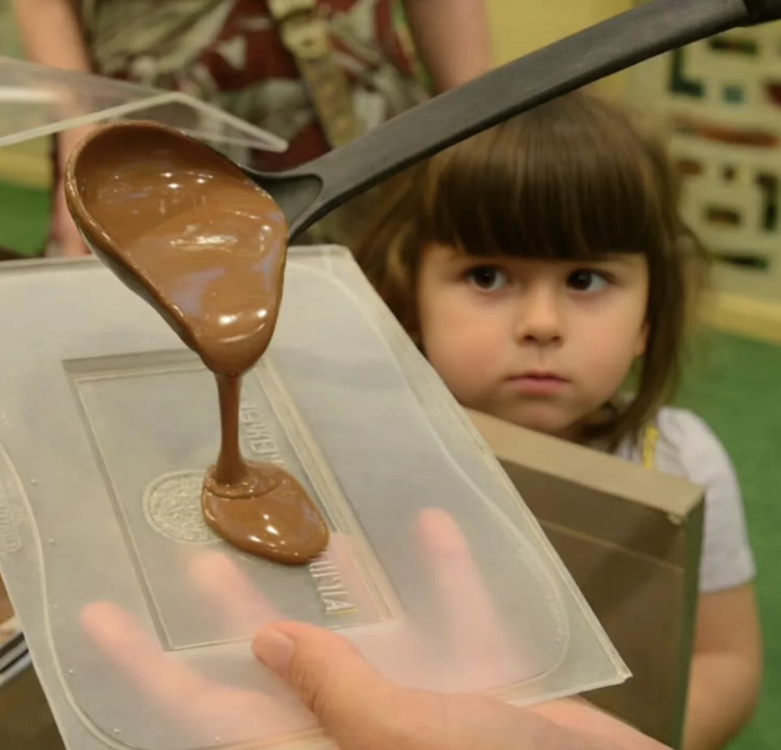 Close de uma concha derramando chocolate derretido em um molde enquanto uma menina observa atentamente.