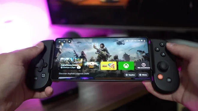Uma pessoa segurando um smartphone acoplado a um controle de jogo portátil, com um jogo de ação na tela e iluminação LED ambiente rosa ao fundo.