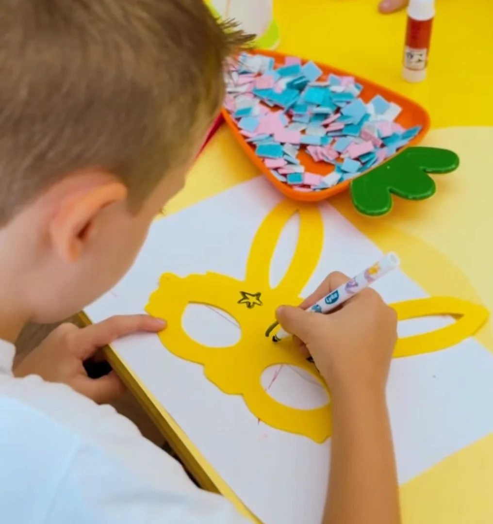 Close-up de uma criança em uma mesa amarela desenhando em uma máscara de coelho amarela.