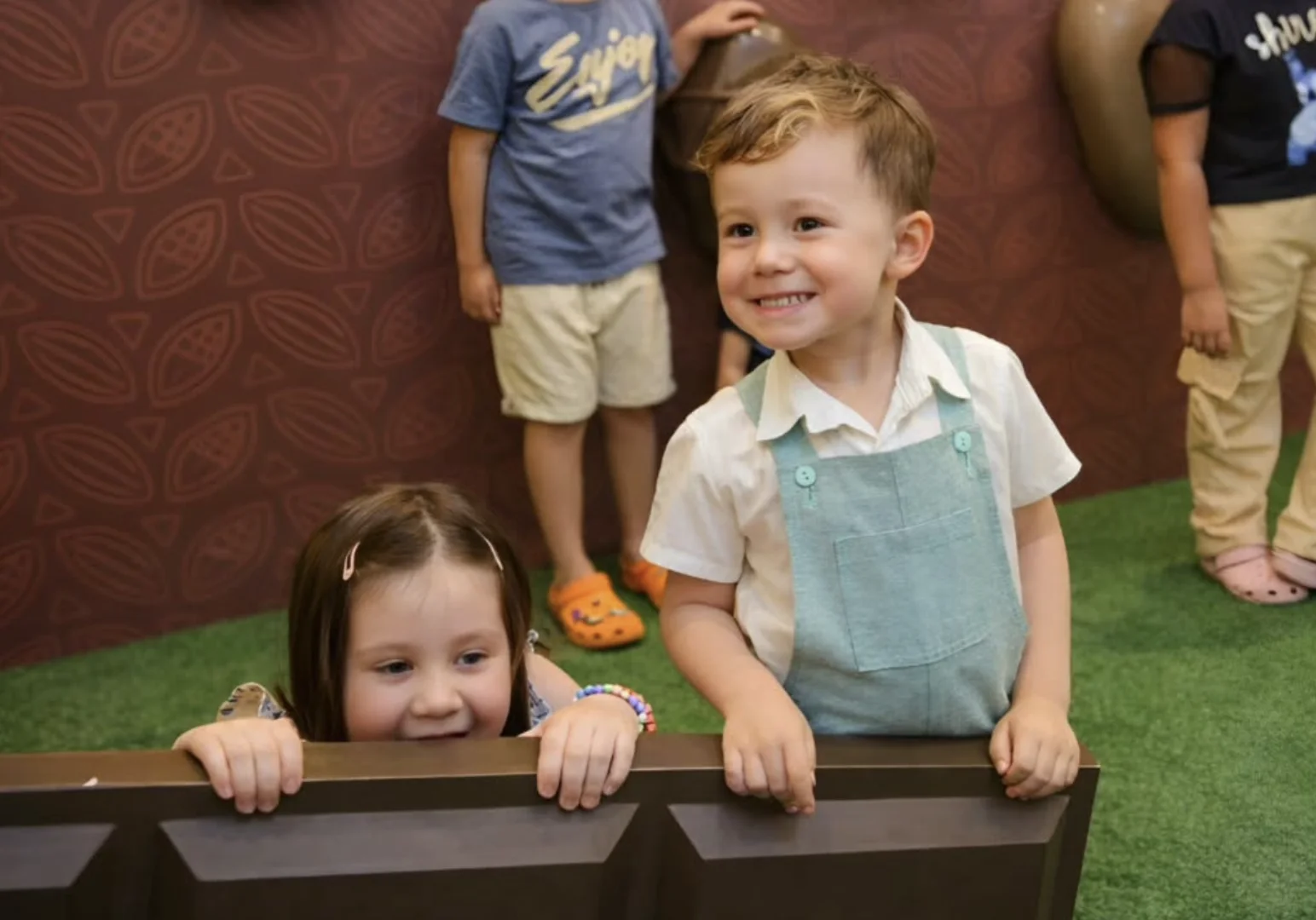 Dois meninos sorridentes em primeiro plano posam em frente a uma parede marrom com padrões.