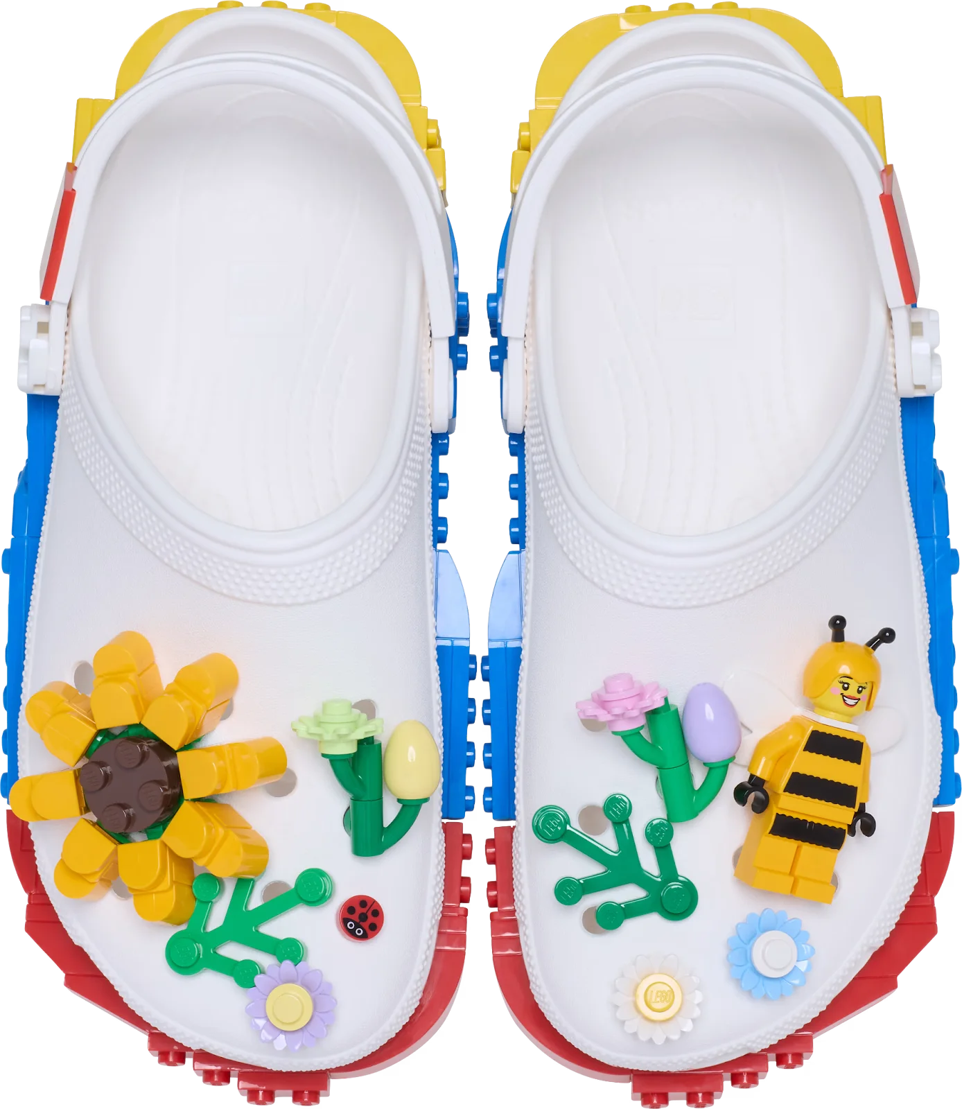 Par de Crocs brancas vistas de cima, decoradas com peças de LEGO coloridas em formato de girassol, abelha, flores e uma joaninha. O solado lateral apresenta blocos de LEGO em azul, amarelo e vermelho.